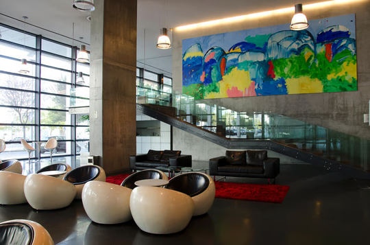 Modern lobby with egg-shaped chairs, black sofas on a red carpet, and a vibrant abstract painting. Glass wall and stairs create an open, stylish atmosphere.