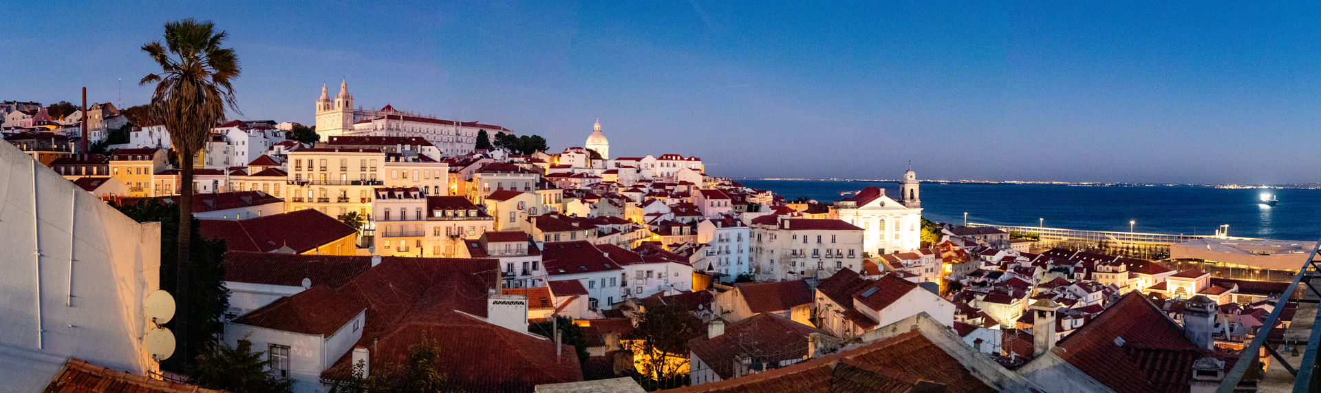 Panoramic view of Lisbon's Alfama district at dusk. Warm lights illuminate red-roofed buildings and historic churches.